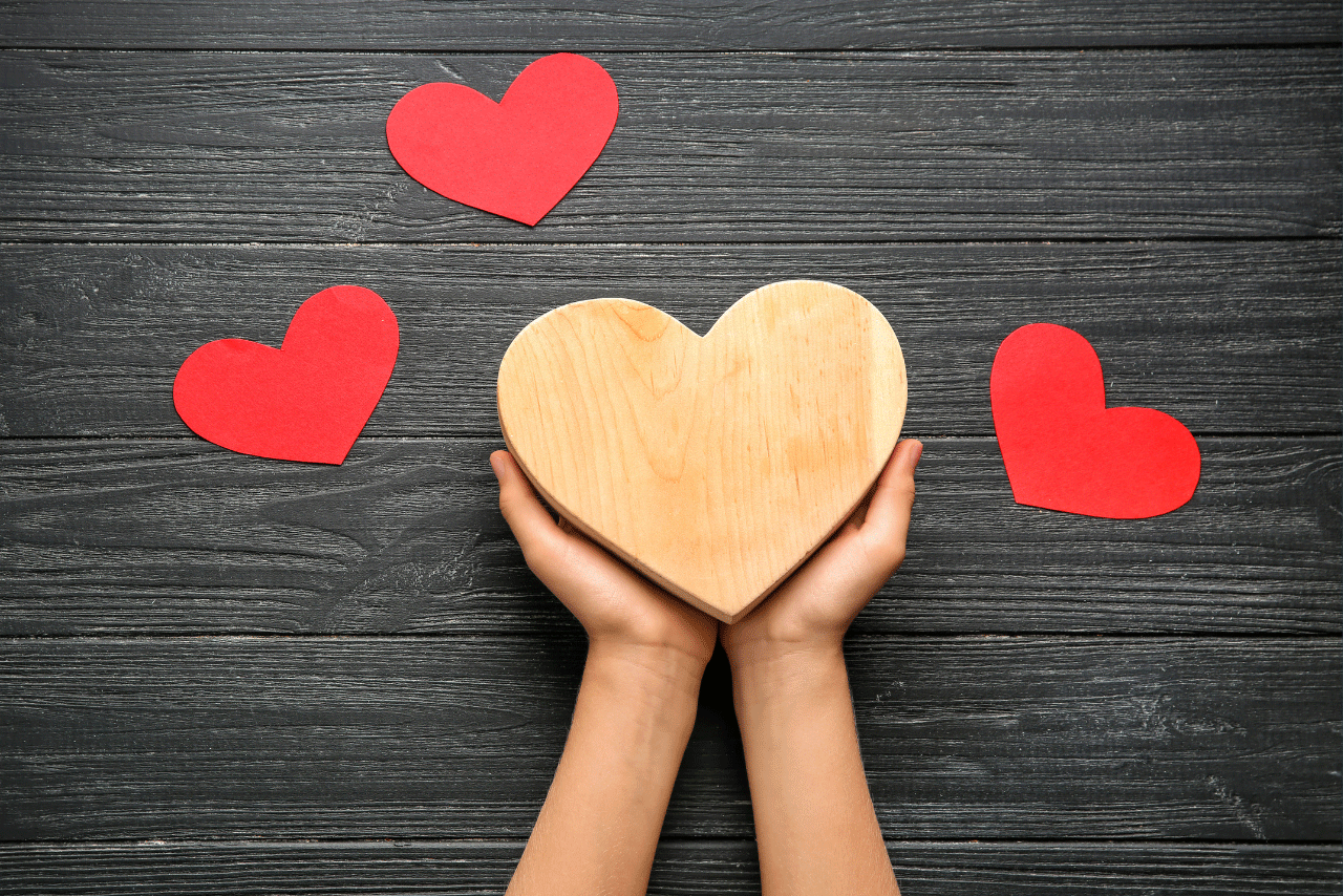 Hands holding a wooden heart with three red paper hearts around them. 