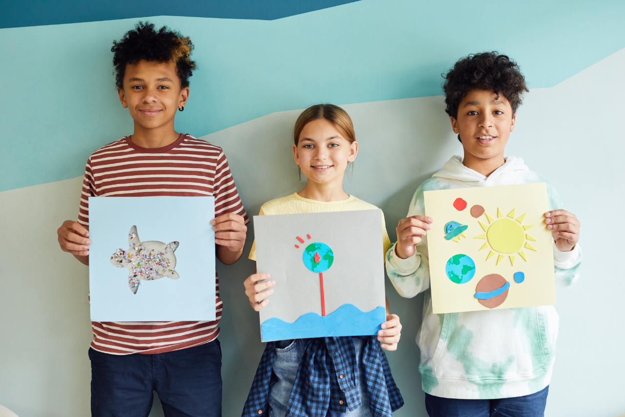 Two boys and a girl standing up against a wall holding artwork they created.