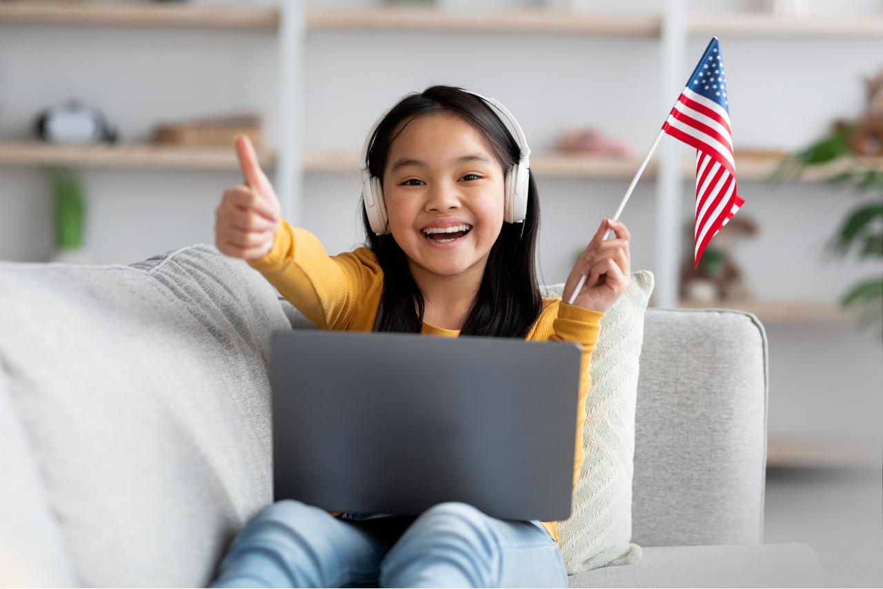 Girl sitting on a couch with a laptop holding an America Flag giving a thumbs up. 