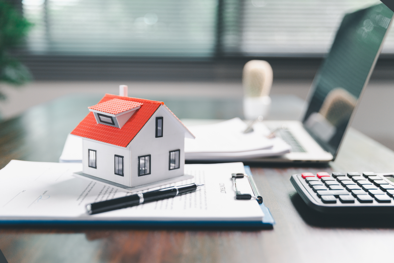 A model house sitting on a clipboard next to a pen, laptop, and calculator.