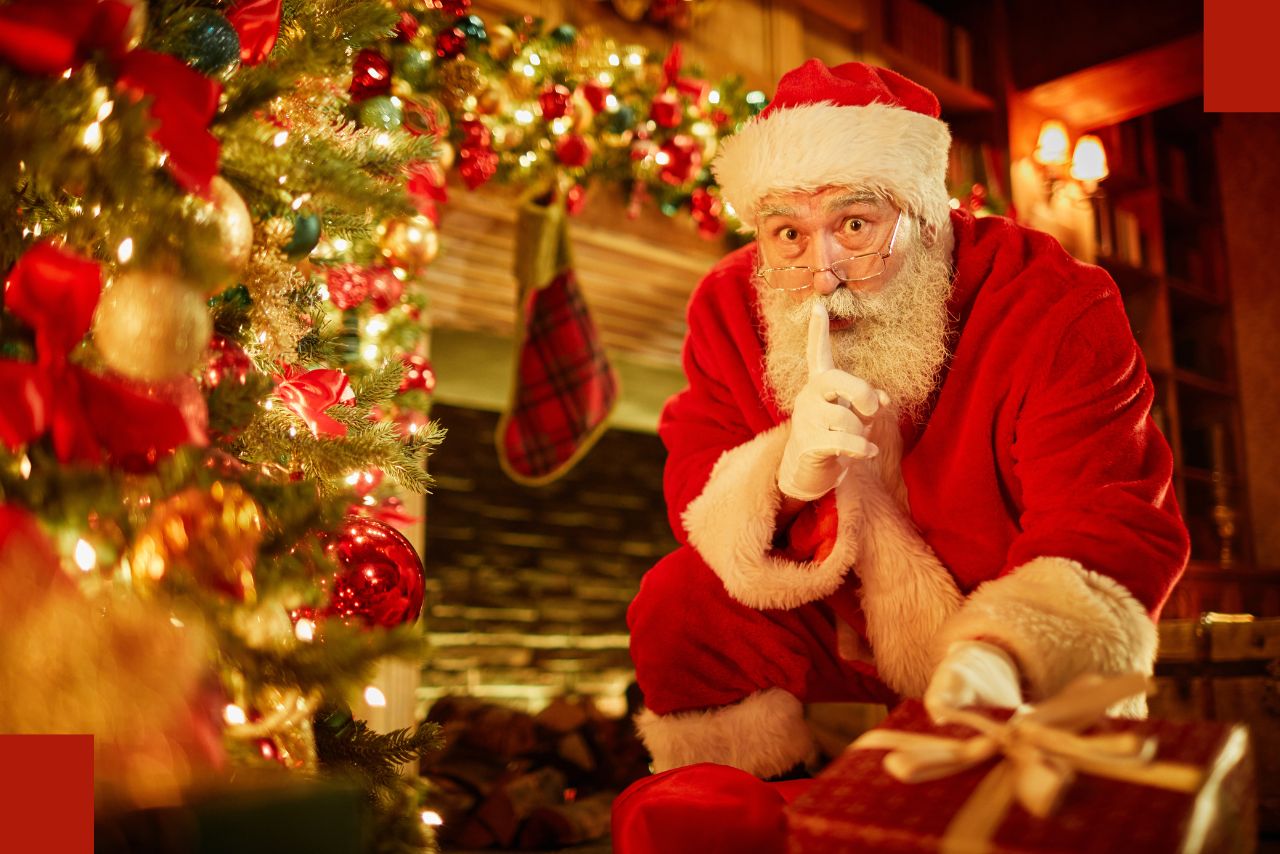 Santa Clause sits by a decorative Christmas scene by a fireplace with a finger to his mouth in a "shhh" gesture.