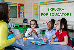 Three little girls sitting at a round table in a classroom looking at their female teacher holding a paper. 