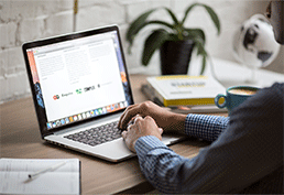 Side profile of person sitting at desk with hands on laptop. Paperwork and a plant sit on the desk to the right of the laptop. 