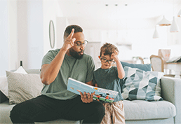 bearded man with glasses reading a book using sign language to a little boy wearing glasses. 