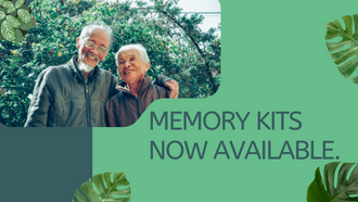 Elderly man with facial hair wearing a jacket and glasses hugging an elderly lady wearing a jacket in from of a blooming bush. Text states memory kits now available as dark green lettering over a light green box. 