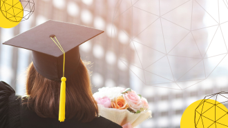 Back of woman wearing a graduation cap and gown. She is holding a bouquet of pink roses looking at office buildings.