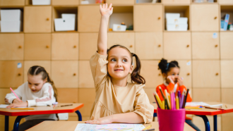 3 girls are sitting at desks in a classroom. One girl has her hand raised, the second is writing on paper, and the last girl is looking out of frame.
