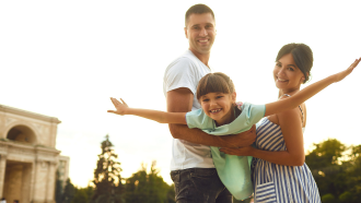 Man in a white t-shirt and jeans facing a woman wearing a sundress. Both are holding a little girl in an airplane pose who is wearing a green dress. 