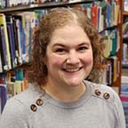 Board Member Kait Gallagher-Wilsterman sitting in front of bookshelf of books