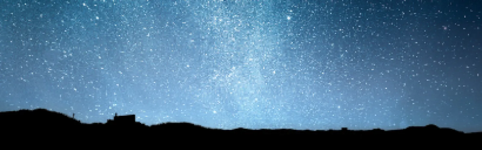 Night sky featuring lots of stars over the shadow of a mountain range. 
