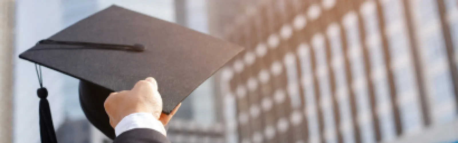 Hand holding a graduation cap in front of office buildings. 